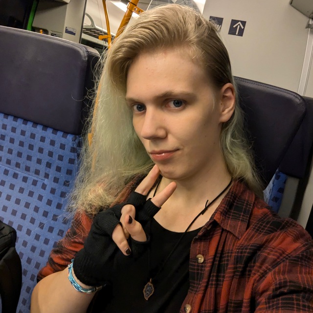 A photo of me sitting in a 2nd class passenger seat on a TALENT 2. I'm looking towards the camera, my blonde/washed-out-blue hair is going down the left side of my face, and I'm wearing a black shirt, black fingerless gloves and a red flannel. I also am wearing a necklace of a glass shard from a Leipzig NGT10 tram.