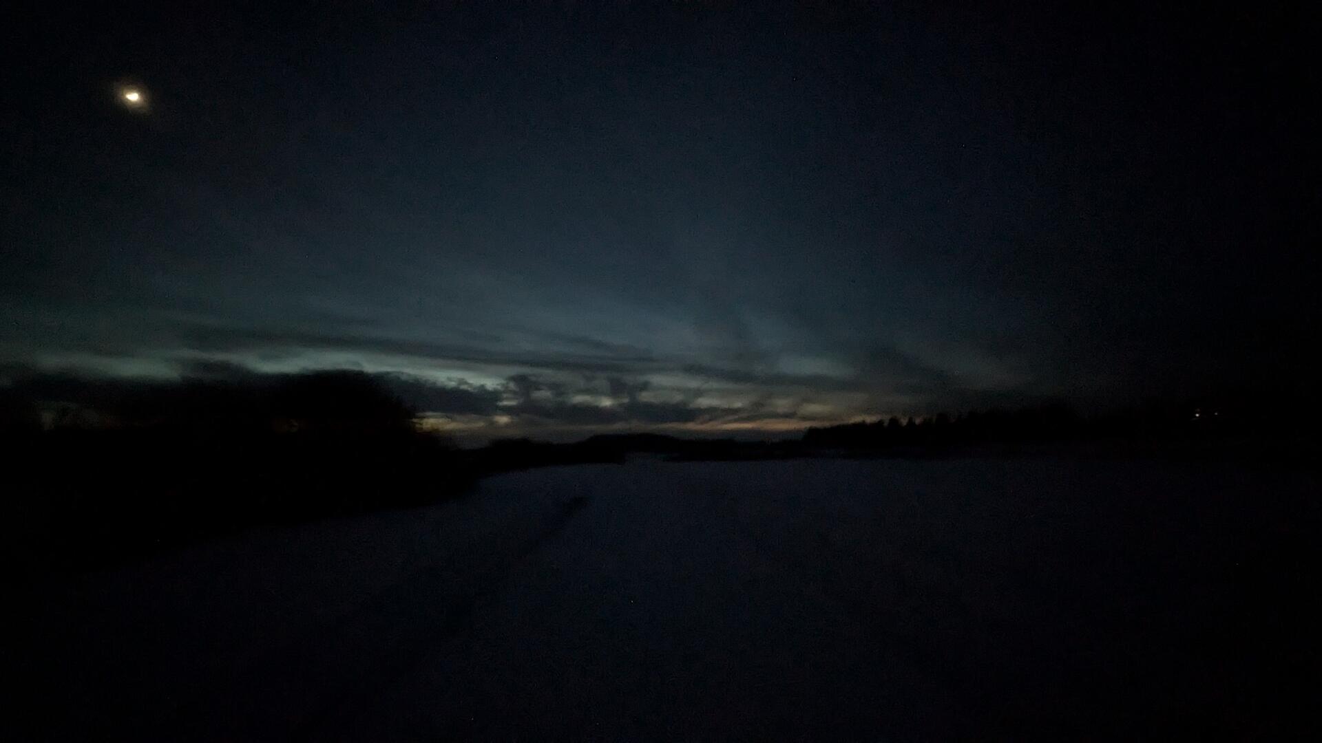 selfsame cross-country skiing tracks, but looking towards the west instead of east. Visible new moon. Very dark blue to black tint, some clouds and some remaining orange and pastel-blue visible from the sun which had already set.