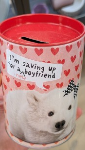 A pink, cylindrical tin coin bank with red hearts on the background and a small polar bear in the foreground printed on it which says 'I'm saving up for a new boyfriend'