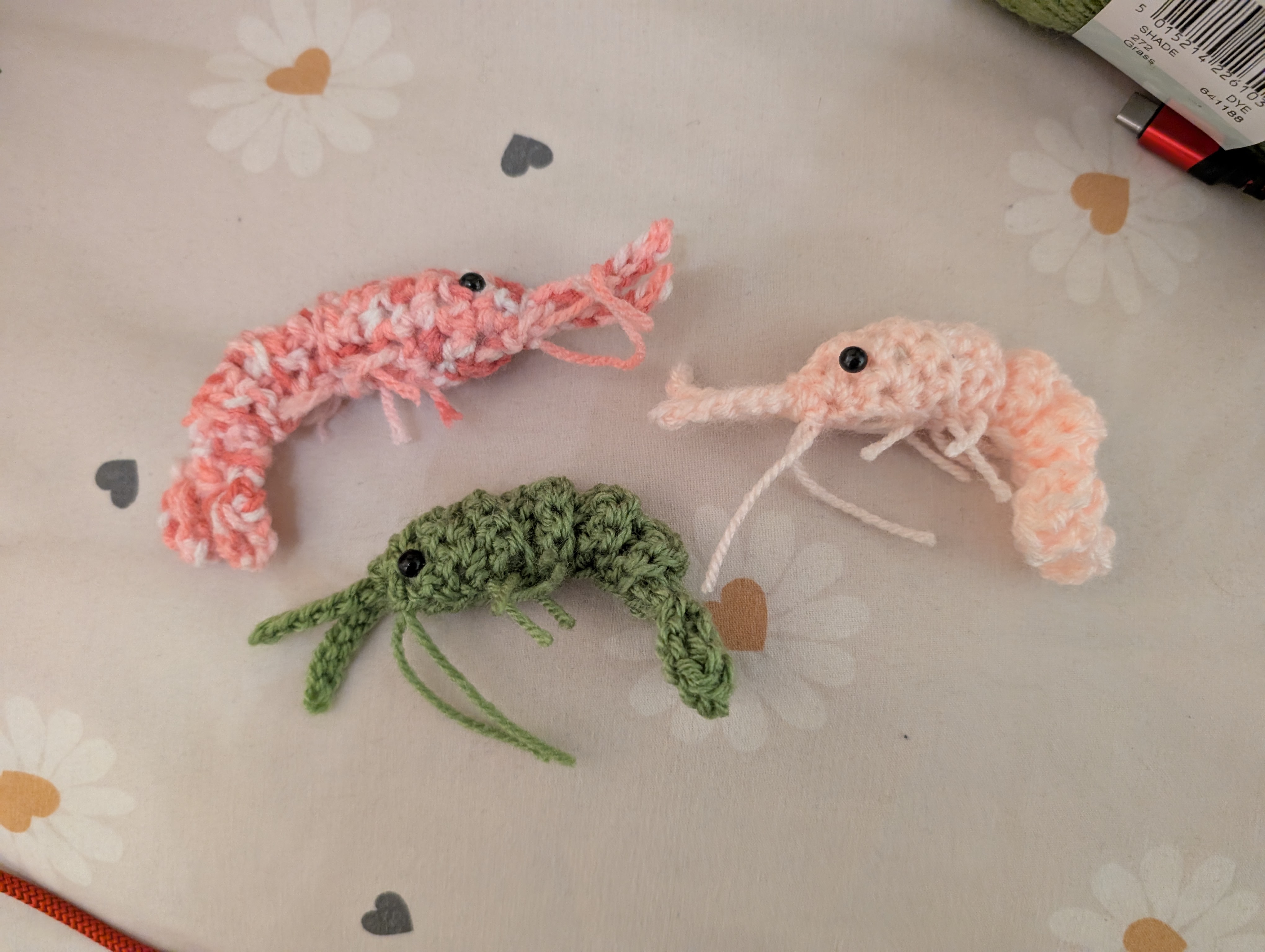 A photo of three small crocheted shrimp - one is a single shade of 
pale pink, one is multi-coloured light & dark pink with flecks of white, 
and one is 
green